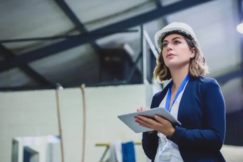 A focused female engineer wearing safety gear while doing an inspection on machinery inside of a workshop